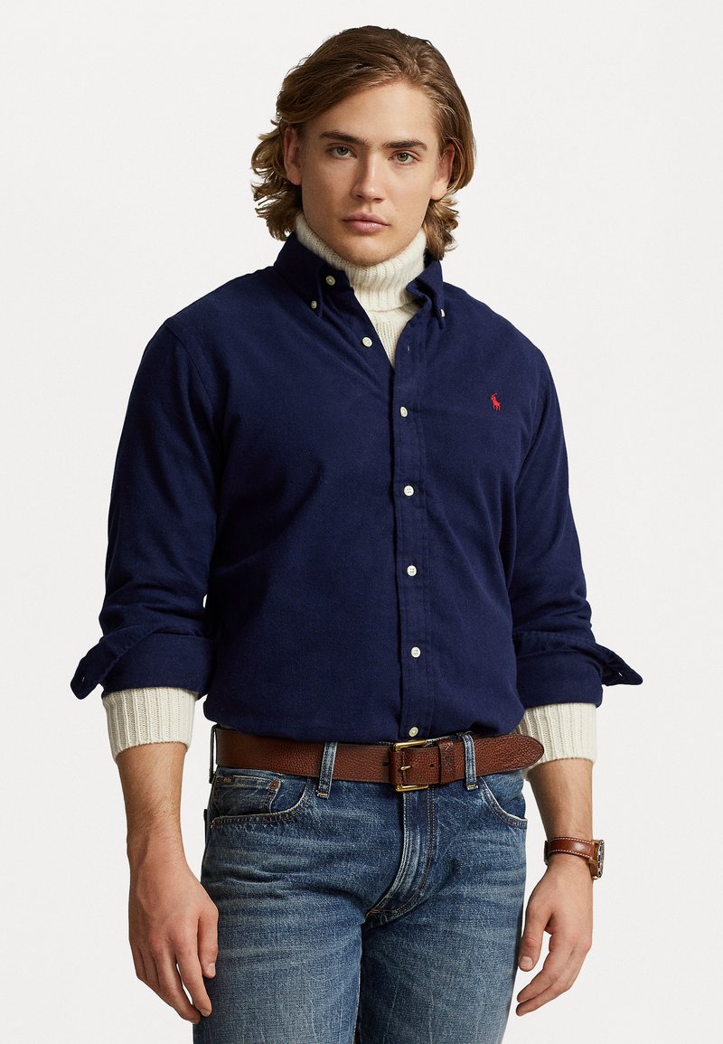 Camisa de algodão azul-escuro com botões brancos, apresentando mangas arregaçadas sobre um suéter creme, acompanhada de um cinto de couro castanho e jeans.