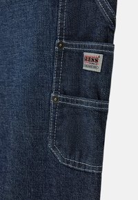 Poche de jean en denim bleu foncé avec double couture blanche, rivets métalliques et une petite étiquette rectangulaire en tissu indiquant « Guess Originals Established 1981 ».