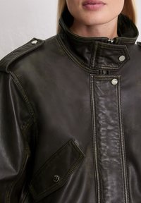 Dunkelolivgrüne Lederjacke mit hohem Kragen, silberner Hardware, Schulterklappen und strukturierten Taschenapplikationen.