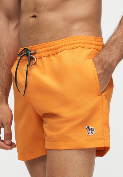 Homme portant un short de bain orange vif avec un cordon noir et bleu et un petit logo zèbre coloré sur la jambe gauche.