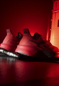 Paire de baskets Adidas Ultraboost rouges sur un sol sombre éclairé par une lumière rouge, avec une barre métallique devant.