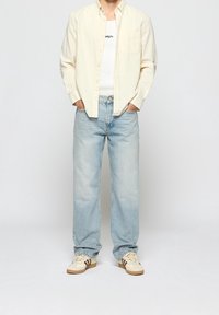 Chemise beige clair boutonnée par-dessus un débardeur blanc, associée à un jean large bleu clair et des baskets crème avec des accents marron.