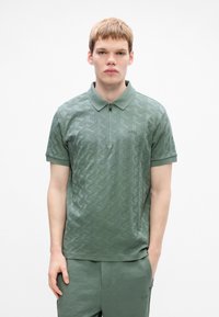 En ung man iklädd en strukturerad ljusgrön polo och matchande byxor står mot en enkel vit bakgrund.