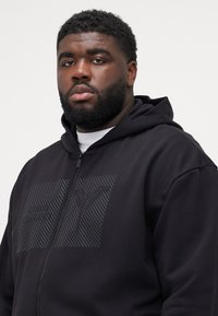 Zwarte zip-up hoodie met een getextureerd logo in een donker patroon. Het materiaal lijkt zacht met een relaxte pasvorm en een gelaagde capuchon.