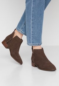 Osoba ve středně dlouhých hnědých sukních botách s nízkými blokovými podpatky a zkrácených modrých denimových džínách na bílem povrchu.