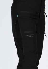 Pantaloni outdoor neri Swedemount con tasca con zip sulla coscia e piccolo dettaglio della bandiera svedese su sfondo bianco.