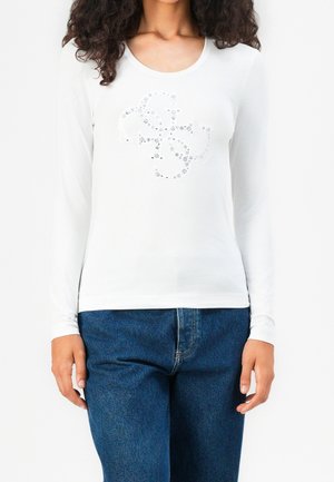 Femme portant une chemise blanche à manches longues avec un motif discret en strass et un jean en denim bleu sur un fond uni.