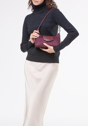 Borsa in suede bordeaux con un design strutturato e chiusura a flap, tenuta da una persona che indossa un dolcevita nero a coste e una gonna crema.
