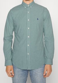 Green and white checkered button-up shirt with a collar, long sleeves, and a small blue logo on the chest. Worn with blue jeans.