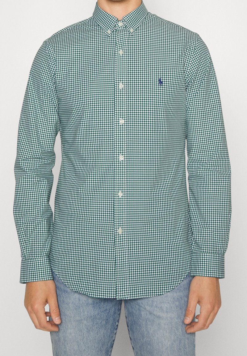 Green and white checkered button-up shirt with a collar, long sleeves, and a small blue logo on the chest. Worn with blue jeans.