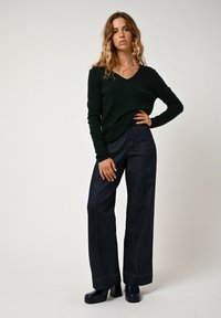 Dunkelgrüner V-Ausschnitt-Pullover aus weichem Material, kombiniert mit hoch taillierten, weit geschnittenen blauen Jeans mit auffälligen Nähten und Taschen.