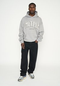 Sudadera gris con la marca "MLTPLY", bolsillo tipo canguro, combinada con jeans negros y zapatillas con estampado. Tejido suave y corte relajado.