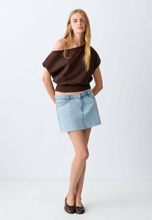 Young woman with long blonde hair wears off-shoulder brown top, light blue denim mini skirt, and brown flats, standing against white background.