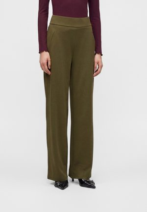 Femme portant un pantalon large vert olive, un haut à manches longues bordeaux et des talons noirs à bout pointu, debout devant un fond uni.
