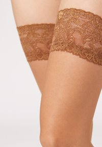 Braune Spitzenstrümpfe bis zum Oberschenkel mit zarten floralen Mustern und einer glatten Textur. Die Spitzenoble halten diskret an der Haut.