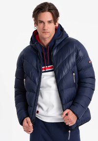 Mörkblå pufferjacka med ett quiltat mönster, huva och dragkedjefickor. Lagd över en vit och mörkblå randig sweatshirt.