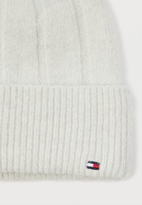 Balta megzta kepurė su įridenti kraštu ir raukšlėtu tekstūru. Maža logotipo užuominos raudonos, baltos ir mėlynos spalvų ant šono.