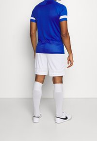 Atleta masculino vestido con una camiseta de manga corta azul, pantalones cortos blancos, calcetas altas blancas y zapatillas Nike blancas, de pie de espaldas.