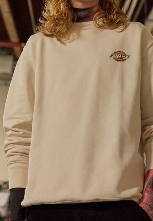 Osoba nosząca kremową bluzę Dickies na kolorowej koszuli z długim rękawem, widoczna od szyi do talii w pomieszczeniu.