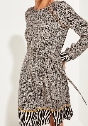Femme portant une robe mi-longue à manches longues avec de petits pois noirs et une bordure à imprimé zèbre aux poignets et à l’ourlet, ainsi qu’une ceinture nouée à la taille.