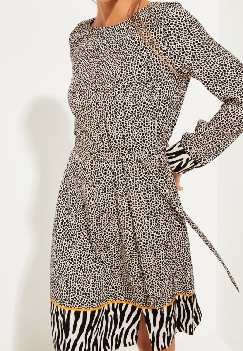 Femme portant une robe mi-longue à manches longues avec de petits pois noirs et une bordure à imprimé zèbre aux poignets et à l’ourlet, ainsi qu’une ceinture nouée à la taille.