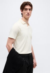 Νεαρός άνδρας που φοράει κρεμ κοντομάνικο πόλο, μαύρο παντελόνι με ρίγες και ζώνη, κρατώντας μαύρο πουλόβερ πάνω από τον αριστερό του βραχίονα, μπροστά σε λευκό φόντο.