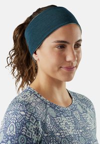 Femme avec des cheveux bruns en queue de cheval portant un bandeau bleu foncé et une chemise à motifs bleus, regardant légèrement vers la droite.