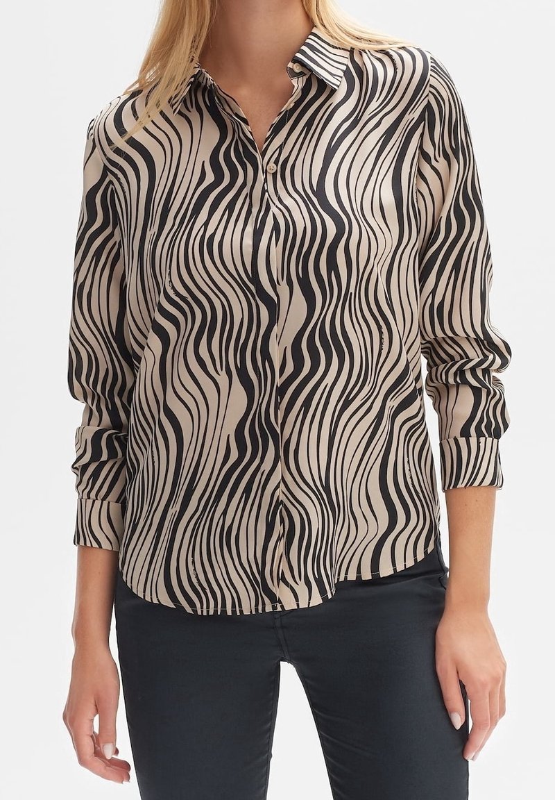 Femme portant une chemise beige boutonnée avec des rayures verticales noires ondulées et un pantalon noir, manches retroussées jusqu'aux avant-bras.