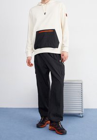 Sudadera blanca con bolsa negra y cremallera naranja, combinada con pantalones cargo negros y zapatillas deportivas negras con detalles en naranja.