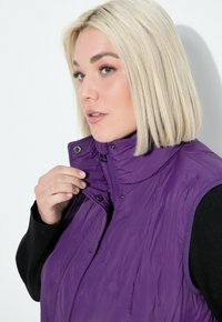 Lila isolierte Weste mit hohem Kragen und Druckknopfverschluss; leichtes Gewebe mit glatter Textur. Das Modell hält den Kragen mit einer Hand fest.
