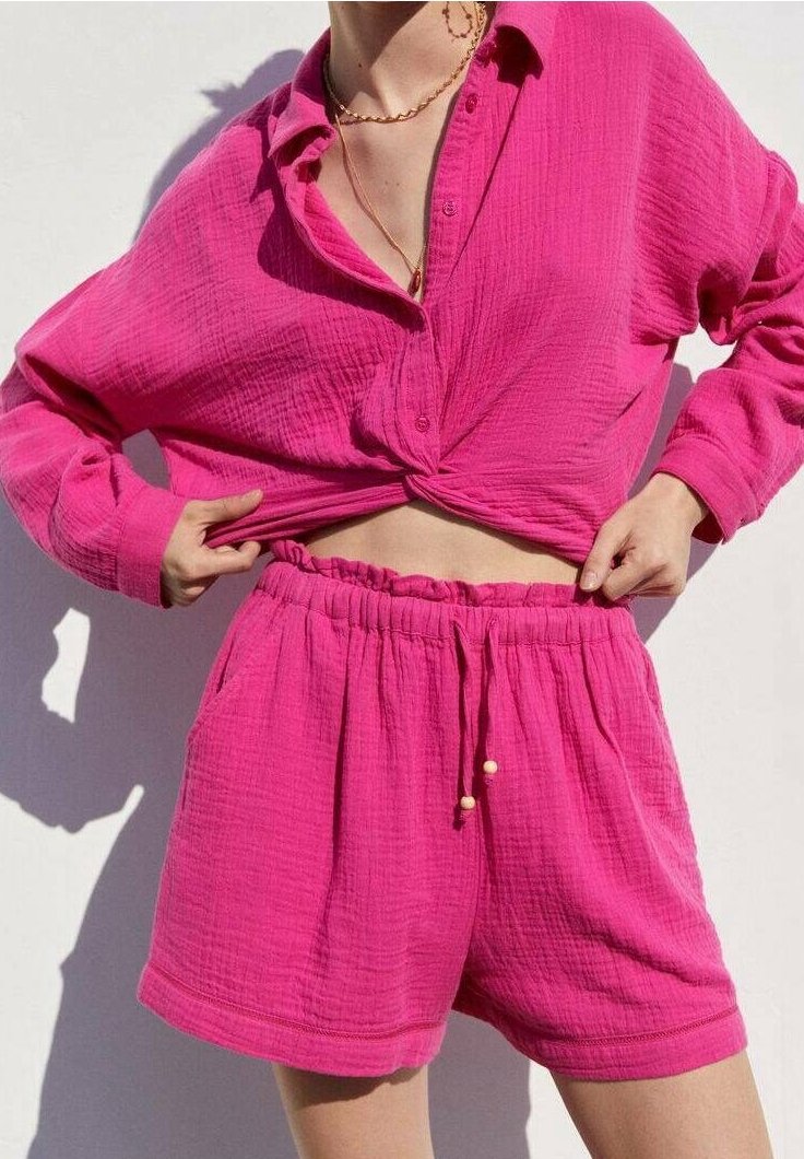 Personne portant une chemise texturée rose vif boutonnée nouée à la taille et un short assorti à taille élastique, sur un fond blanc uni.