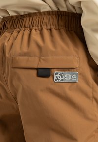 Brune shorts met een elastische tailleband, voorzien van een achterzak en een rechthoekige grijze logo patch met "94." Glad, waterafstotend materiaal.