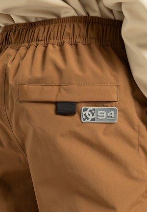 Braune Shorts mit elastischem Bund, ausgestattet mit einer Gesäßtasche und einem rechteckigen grauen Logopatch mit "94". Glattes, wasserabweisendes Material.