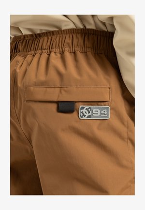 Braune Shorts mit elastischem Bund, ausgestattet mit einer Gesäßtasche und einem rechteckigen grauen Logopatch mit "94". Glattes, wasserabweisendes Material.