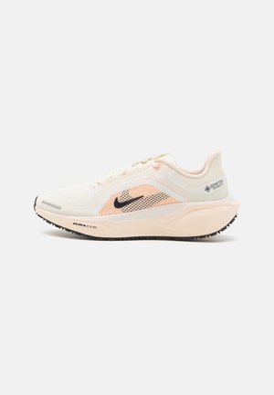 Chaussure de course Nike beige clair avec swoosh noir, panneau latéral en mesh pêche, semelle épaisse avec amorti Air Zoom, et étiquette imperméable Gore-Tex.