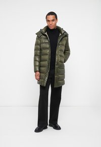 Giacca a vento color verde oliva con chiusura a zip, cappuccio regolabile, polsini a costine e texture trapuntata, abbinata a pantaloni neri e scarpe.