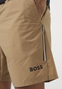 Šviesiai rudos šortai, pagaminti iš lygaus audinio, su juodai baltu brūkšniu šone ir ryškiu juodu "BOSS" logotipu.