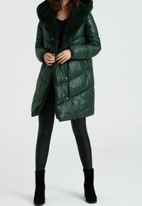 Abrigo de plumas verde con capucha forrada de piel texturizada, acabado brillante, botones a presión y patrón de acolchado diagonal. Llevado con leggings negros y botas.