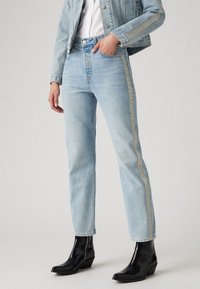 Světle modré džíny s rovnými nohavicemi a metalickou stříbrnou boční lištou, spárované s černými lesklými kotníkovými botami. Obsahují klasický design s pěti kapsami.