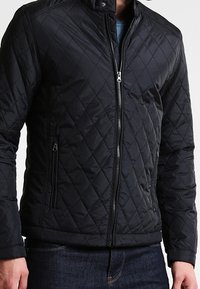 Mand iført en sort quilted zip-up jakke over en blå skjorte med mørke jeans, hænderne afslappet ved siden af.