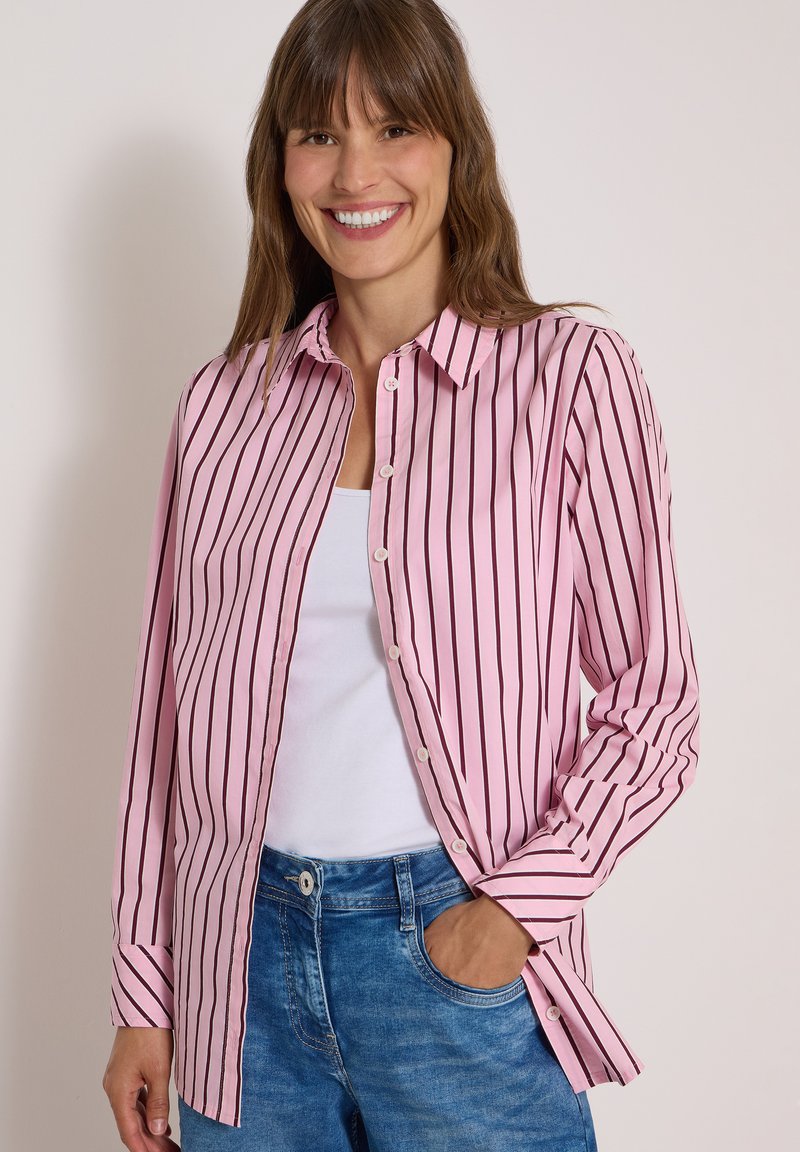 Femme souriante aux cheveux bruns porte une chemise rayée verticale rose et noire boutonnée sur un top blanc et un jean bleu, une main dans la poche.
