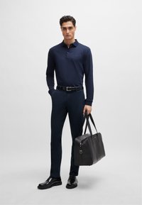 Navy long-sleeve polo, slim-fit dark trousers, polished black shoes, and a black leather tote bag. Clean lines, minimal branding.