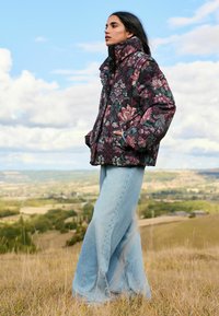 Puffa bunda s kvetinovým vzorom v tmavých odtieňoch s vysokým golierom a bočnými vreckami, skombinovaná s svetlo modrými širokými džínsami v trávnatom prostredí.