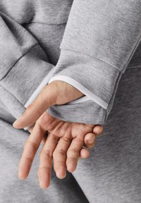 Šedá mikina s texturovanou látkou, se výrazným bílým lemem na manžetě, pod kterou je vidět ruka.