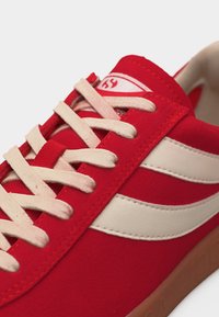 Roter Turnschuh mit weißen Schnürsenkeln, weißen Seitenstreifen und brauner Sohle, Nahaufnahme aus einem oberen Seitenwinkel.