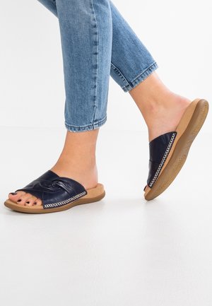 Personne portant des sandales en cuir bleu à enfiler avec des semelles beiges et un jean retroussé aux chevilles, debout sur un sol blanc.