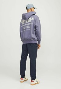 Sudadera morada con estampado blanco en la parte de atrás, combinada con pantalones jogger azul marino. El modelo lleva una gorra azul claro y zapatillas crema con acentos rojos.