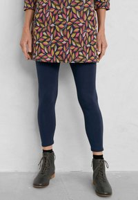 Duge hlače tamnoplave boje, u kombinaciji s haljinom do koljena koja ima šareni uzorak lišća. Cipele su tamnosive s vezicama i drvenim potplatima.