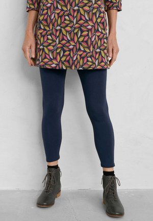 Leggings in marineblauw, gecombineerd met een jurk tot op de knie met een kleurrijk bladerenpatroon. De schoenen zijn donkergrijs met een vetersluiting en houten zolen.