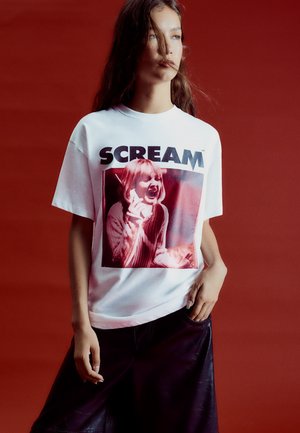 Une jeune femme pose devant un fond rouge, vêtue d’un T-shirt blanc portant le texte « SCREAM » et une image tirée d’un film d’horreur, ainsi que d’une jupe noire.